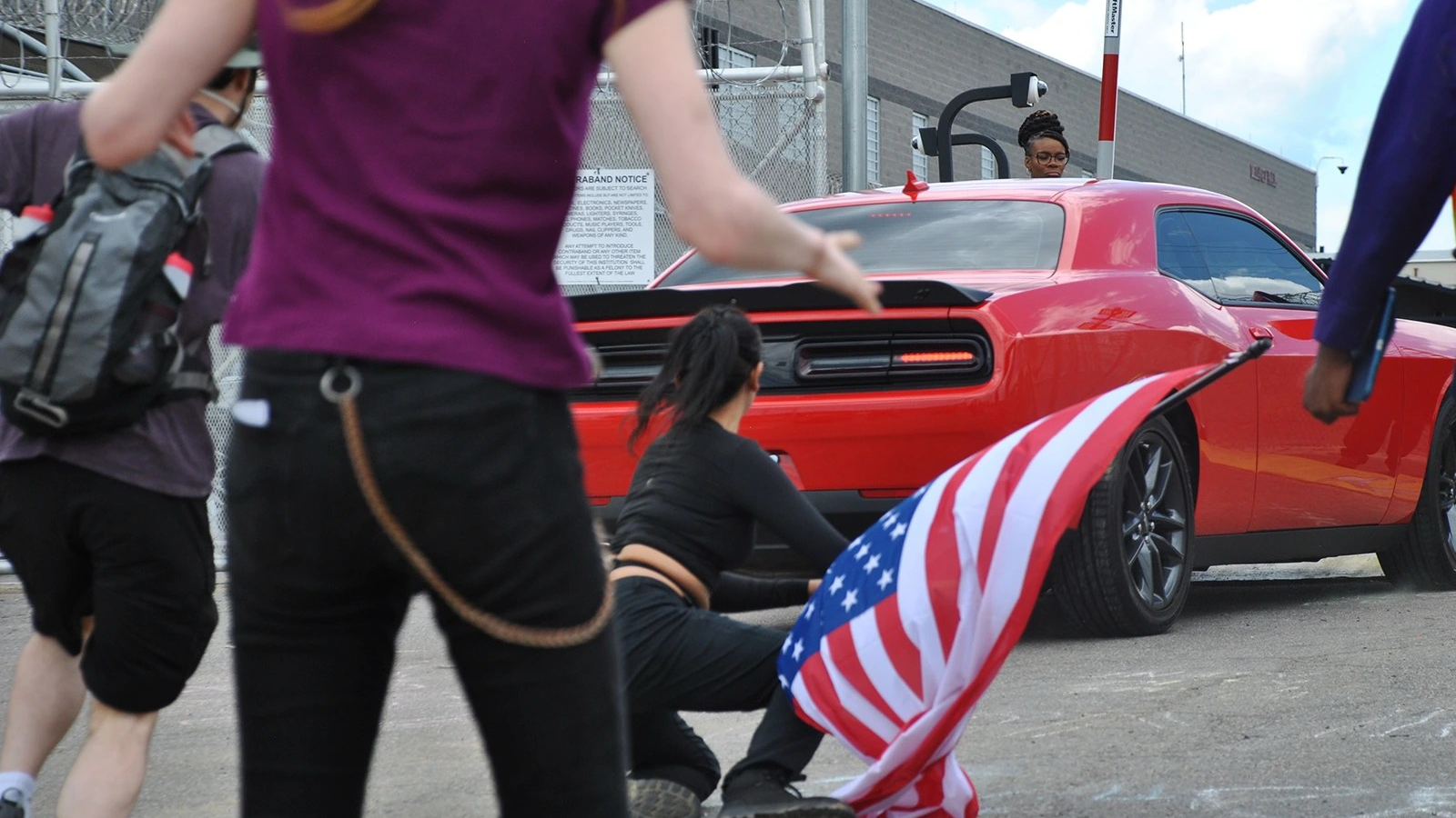 A red car is stopped in front of a gate arm. A woman in all black holding an upside down American flag gets up from the ground. People surround the scene.