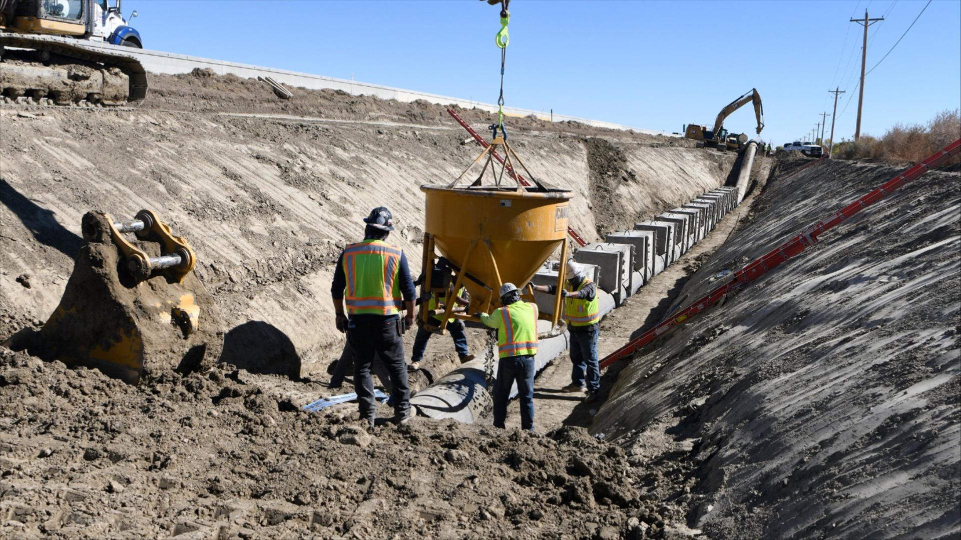 Workers in yellow high-visibility safety garments and hard hats set a concrete bucket on the ground of a construction zone.