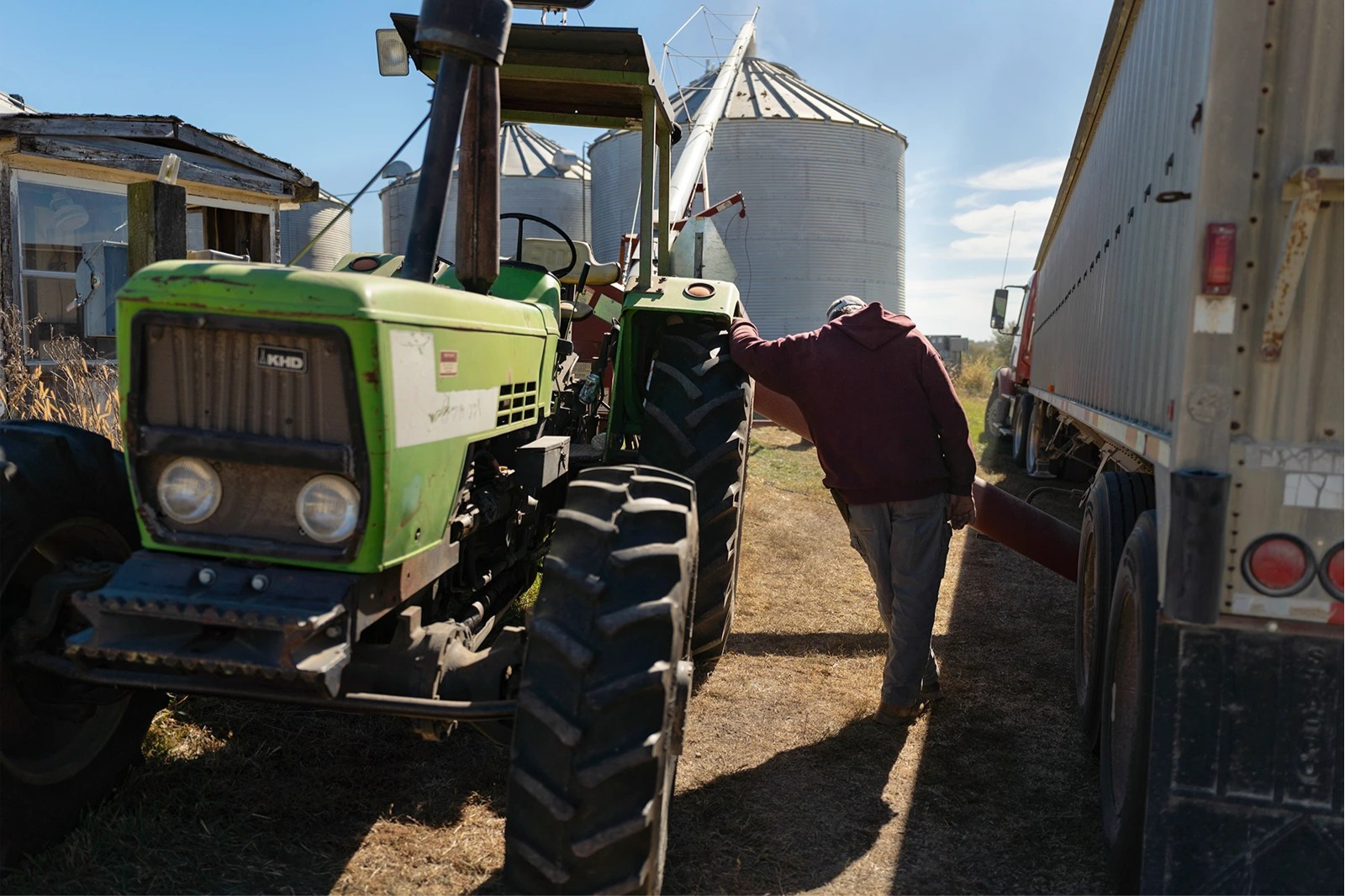 A farmer, with their back to the camera and head down, leans on the wheel of a green tractor on the left of the frame. A grain truck is on the right.