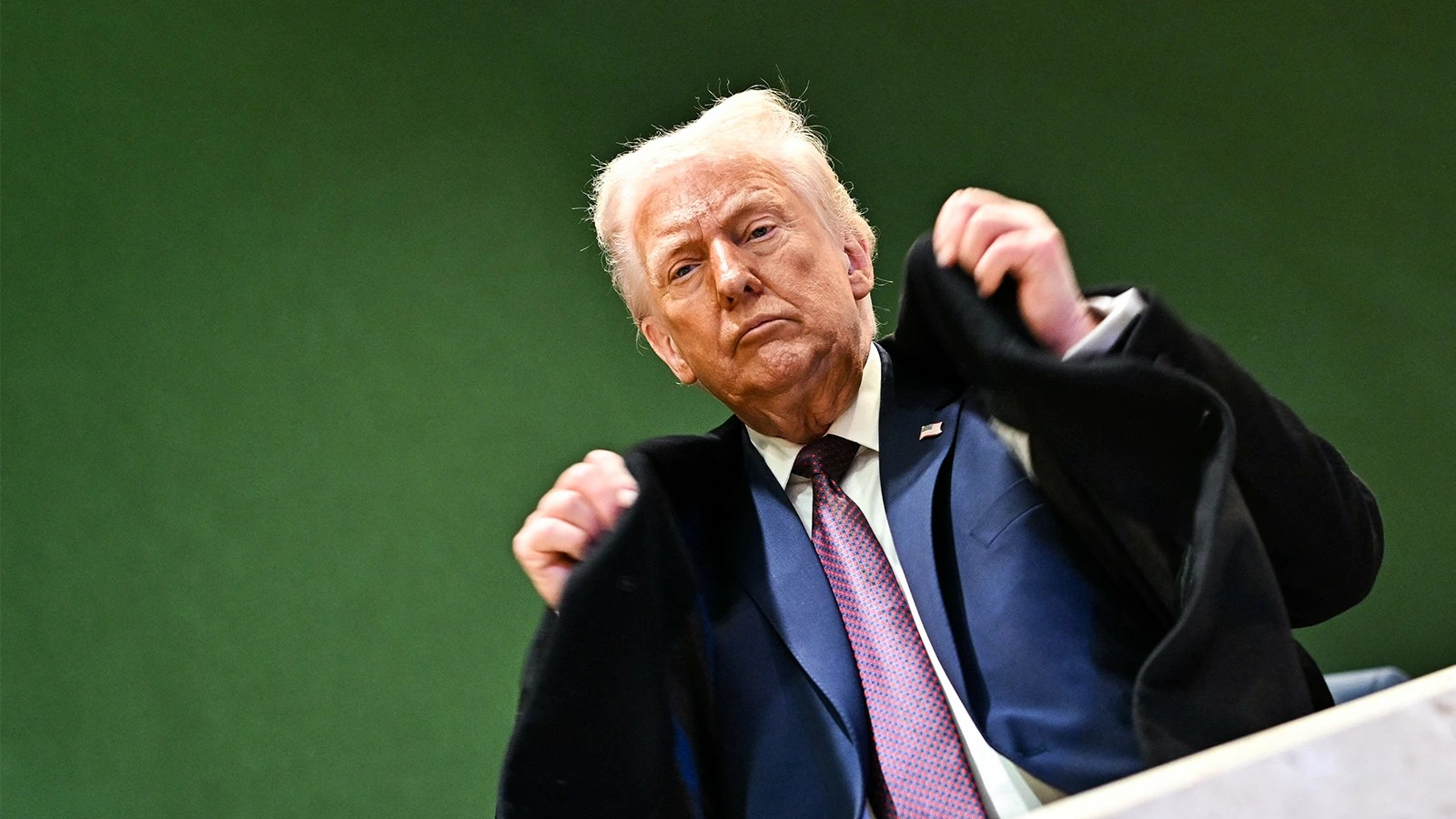Donald Trump adjusts his jacket while standing on a staircase in front of a green wall. The photo is taken from the ground level, looking up at Trump.