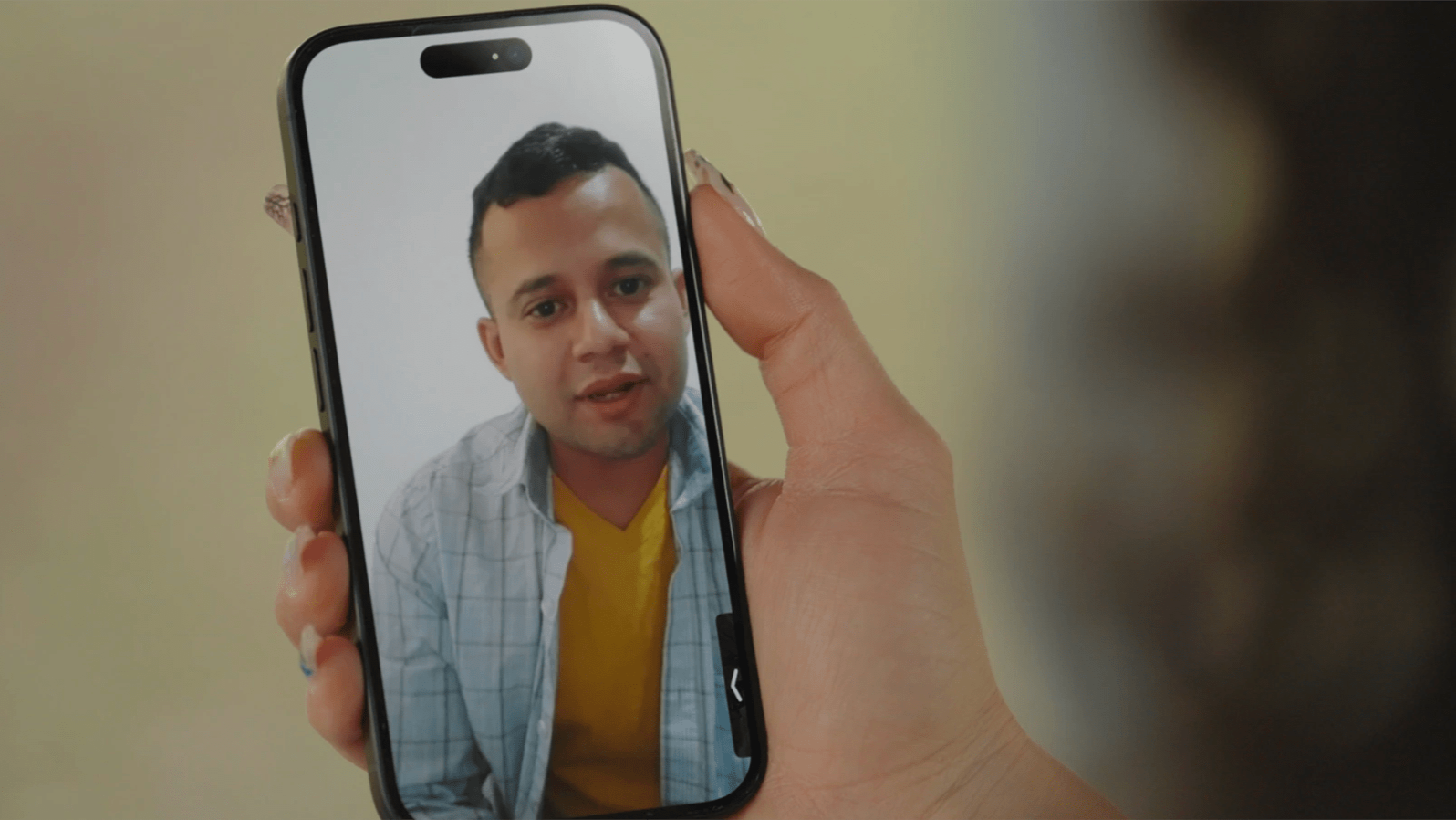 A close up of a woman holding a phone which is video calling with a man in a yellow shirt and blue flannel jacket.