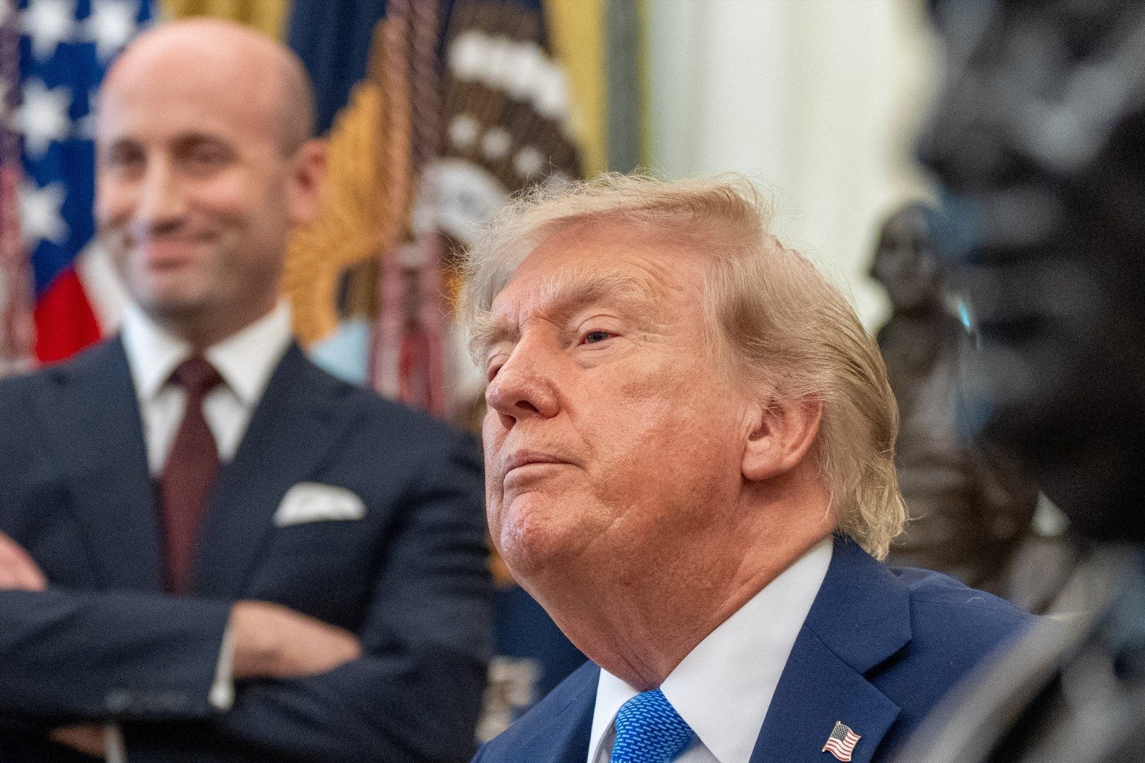 An image of Donald Trump's head in the foreground and Stephen Miller standing with his arms crossed in the background. 