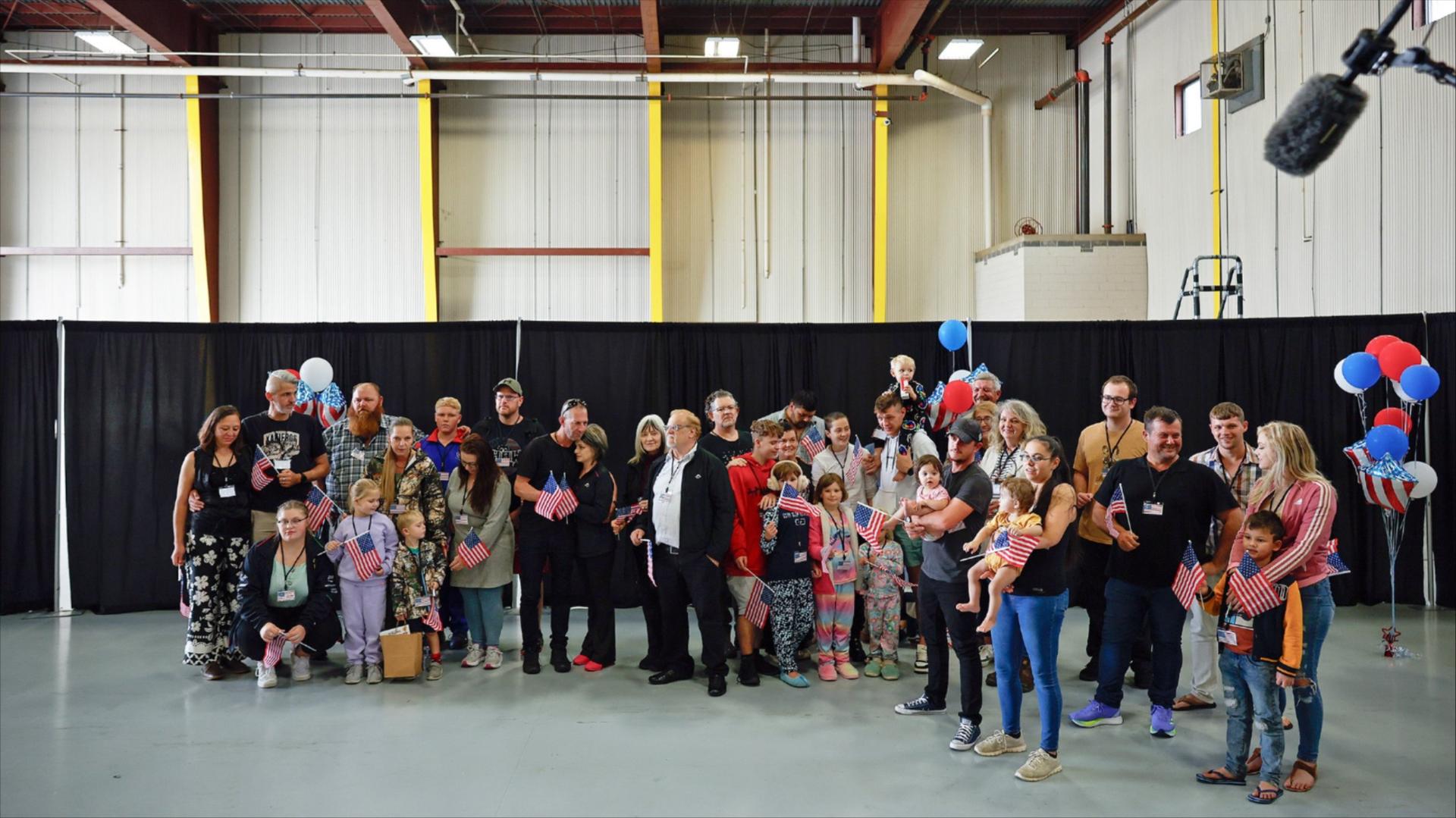 People of all ages stand in an airplane hangar holding American flags