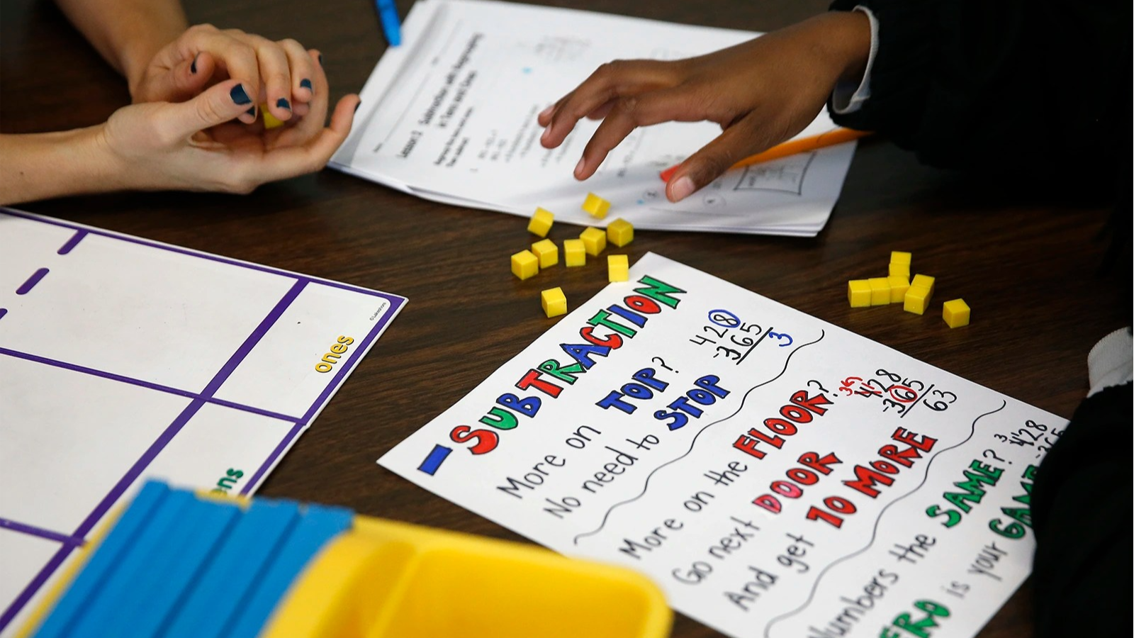 A student's hands and a teacher's hands move yellow counting cubes on a table. A sheet of paper explains subtraction with rhymes.