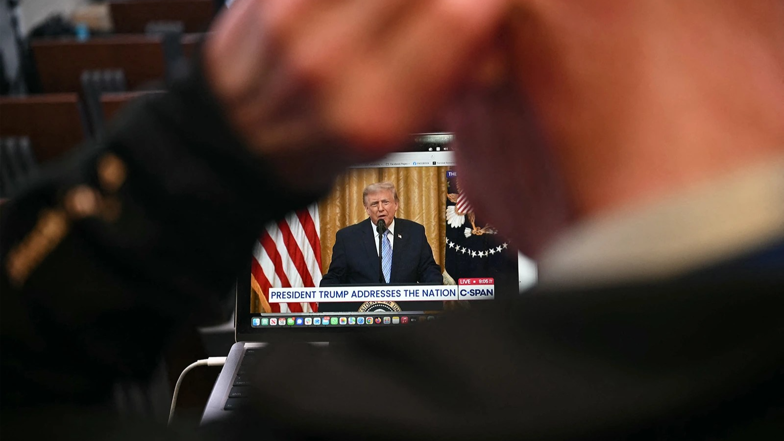 Out of focus in the foreground, a person holds their hand to their ear. In the space between their arm, Donald Trump is seen speaking on a computer.