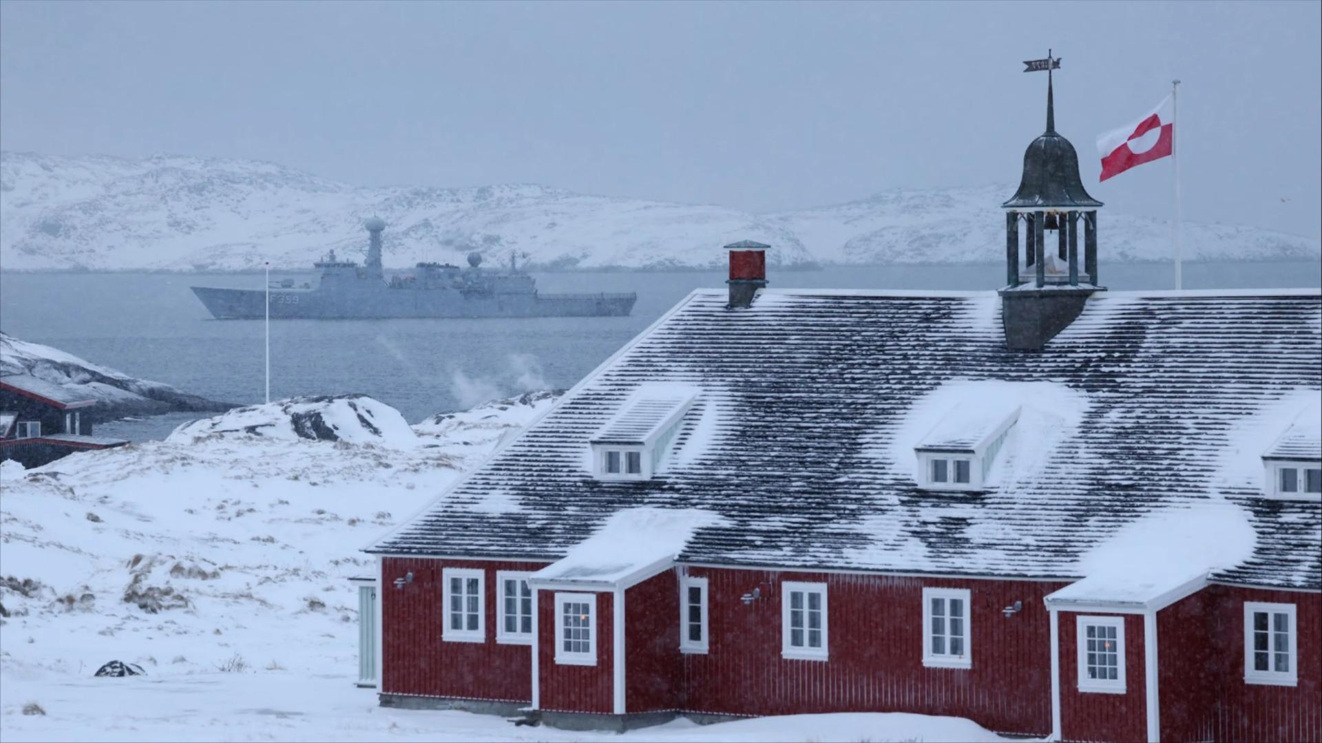 Greenland flag flies over building with Danish Navy ship in background