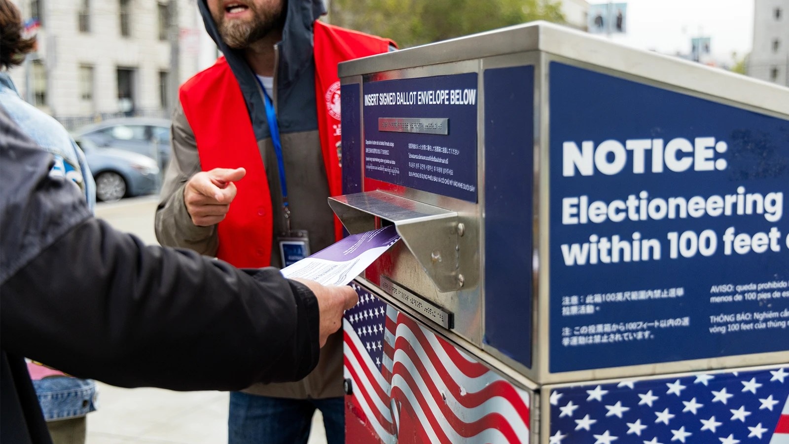 A person's hand reaches into the ballot box form the left side of the frame as another person in a red west talks. Right side of the frame is taken up by the box.