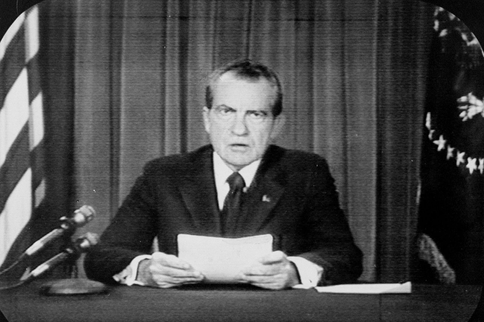 A black and white photo of a television screen. On the screen, an image of Richard Nixon sitting at a table and reading off of paper he his holding with both hands.
