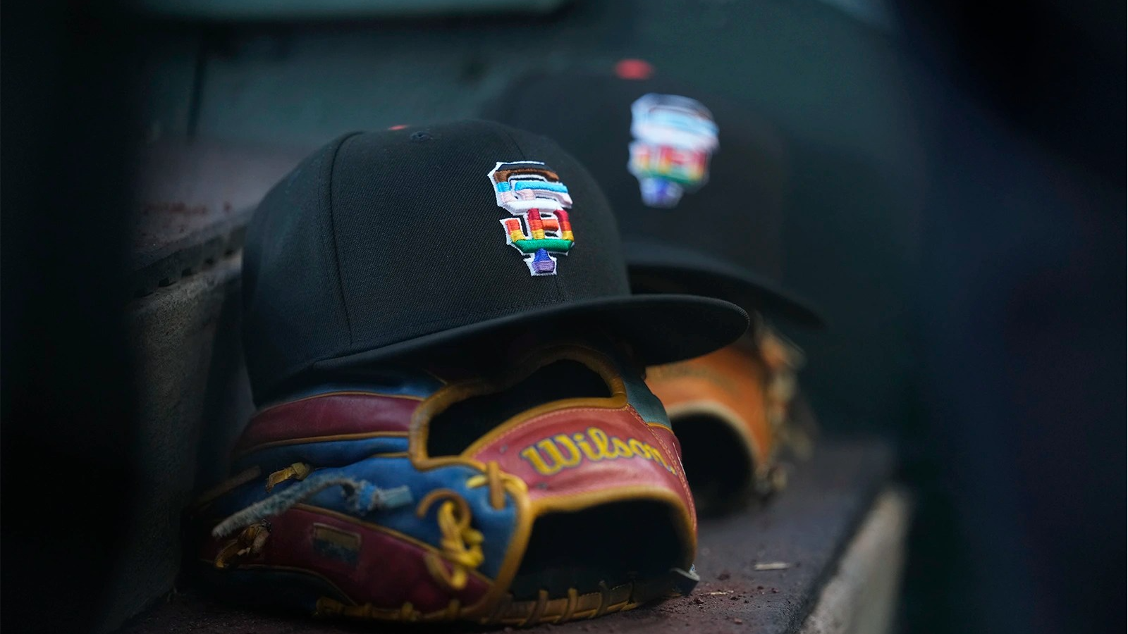 Two baseball gloves, one in front of the other, rest on steps. On top of each glove is a black baseball cap with the San Francisco Giants' logo (an S on top of an F) colored like the progress pride flag.