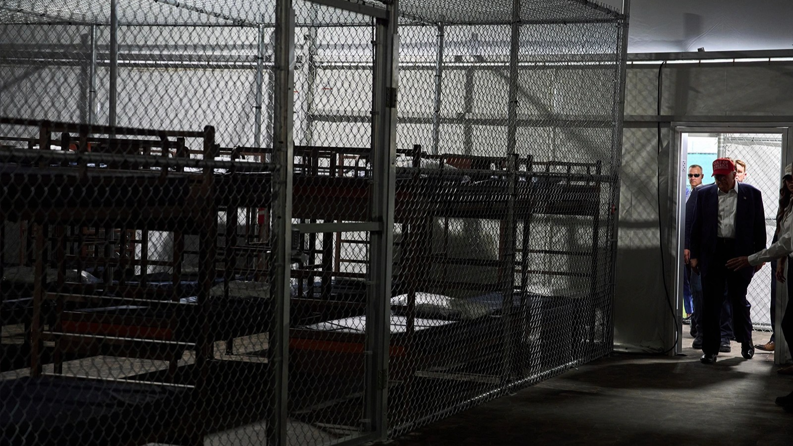 Donald Trump walks through a door on the right side of the frame into a room with many bunk beds in large cages.