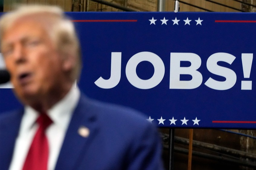Donald Trump speaking into a microphone with a sign behind him