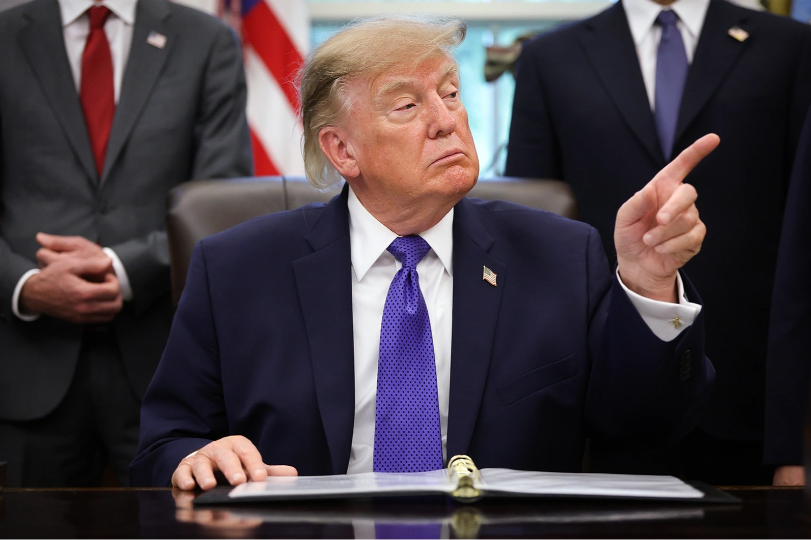 Donald Trump points at something or someone on his left side while sitting at his desk as two men stands behind him.