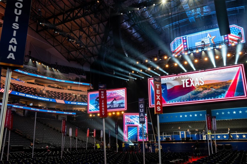 The inside of a large convention center decked out in red, white and blue. Vertical signs on posts say names of states.