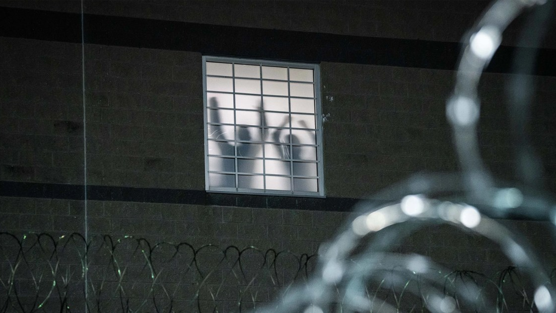 As seen from the outside of Delaney Hall, the silhouettes of two people with their hands up stand behind an opaque window. Barbed wire surrounds the outside of the detention center.