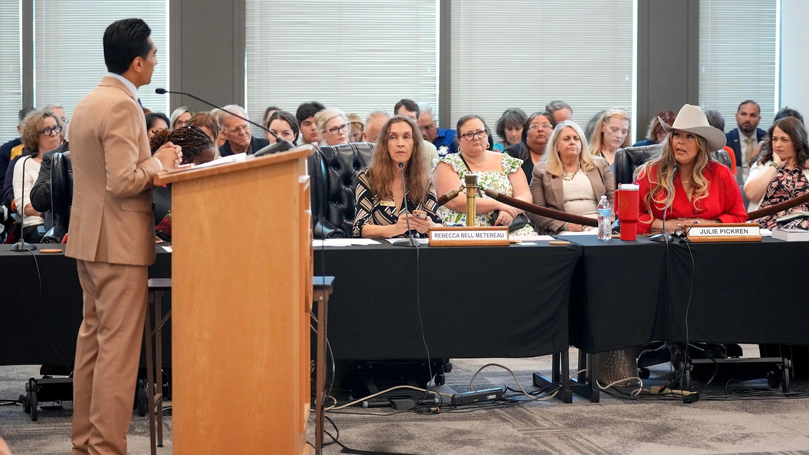 One person in a suit standing at a podium on the left side of the photo looks at a mass of people sitting in the background of the photo.