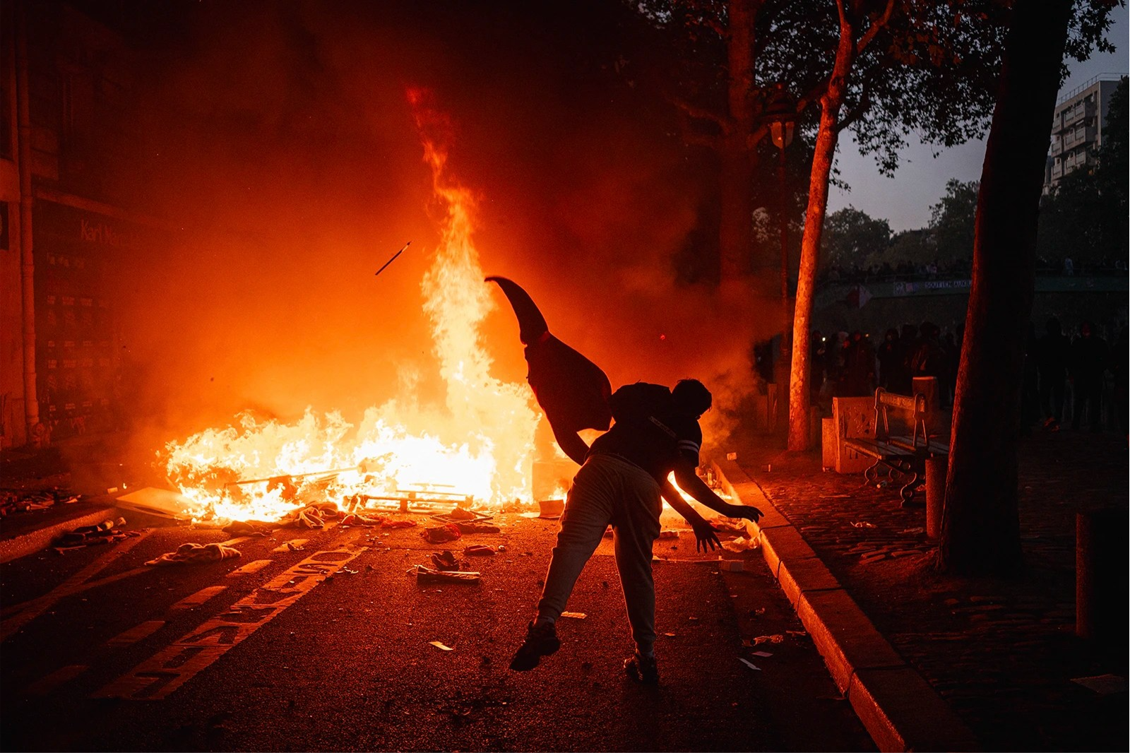 A person with their back to the camera throws something into a large fire on the street.