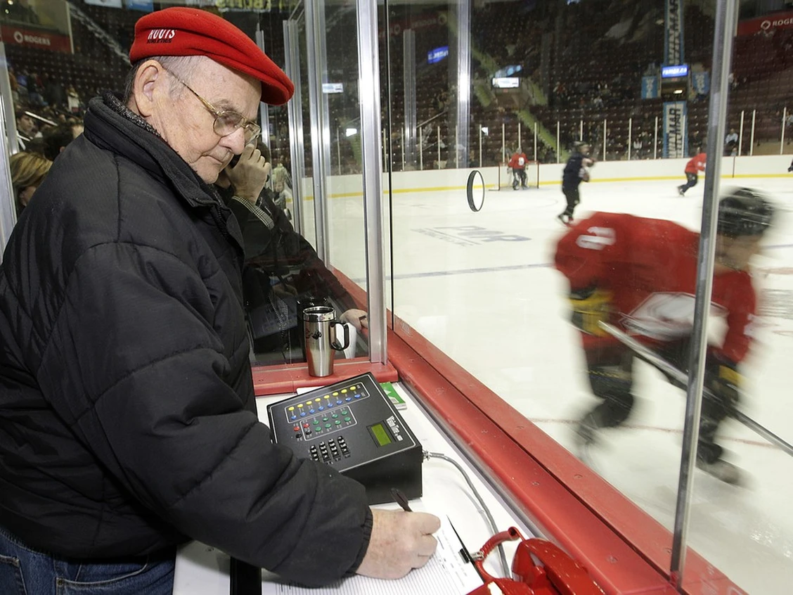 An elderly man in a red hat and a black jacket stands in a timekeeper's booth at a hockey rink.