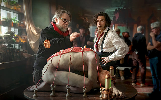 A character standing with their hands on their hips next to a character waving their hand over a person hunched over on a table in a dark room.