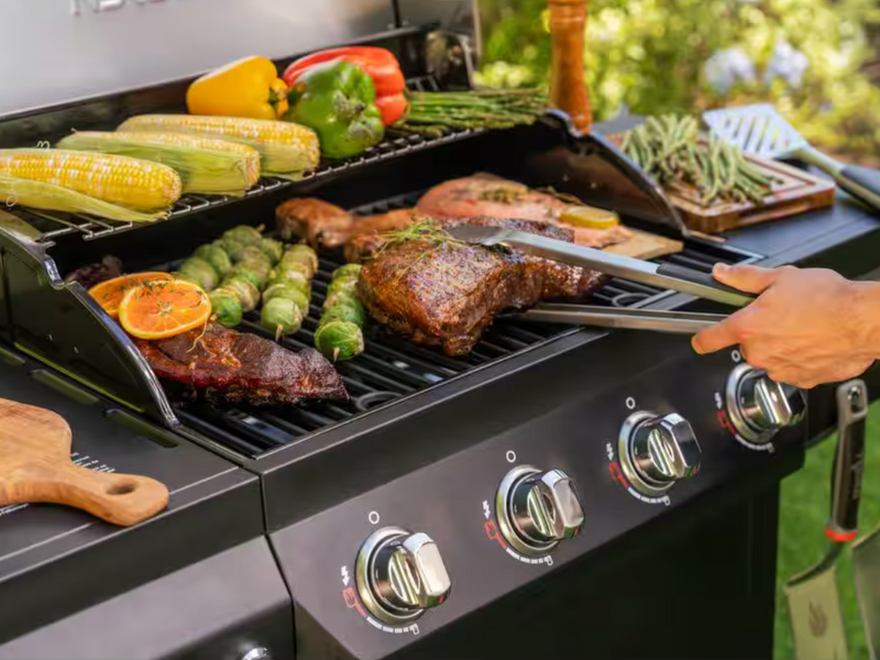 A grill with corn, peppers, and a big steak being turned over by a hand holding a pair of tongs. 