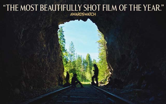Four people hang around train tracks in a dark cave that looks out into a wooded area. Text over reads: 