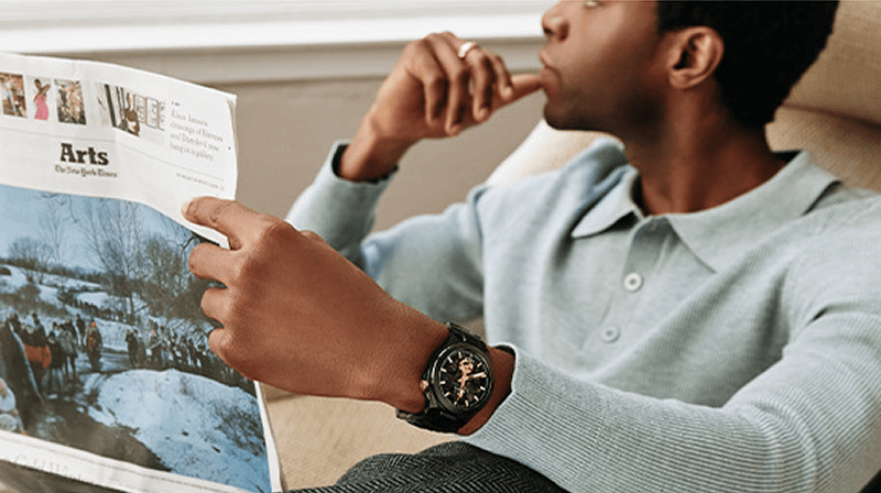 Man reading a newspaper while wearing a black Bulova Maquina automatic watch with gold-tone details.