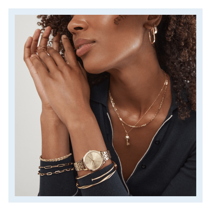 A model wearing layered yellow gold necklaces, bracelets, hoop earrings and a gold watch.