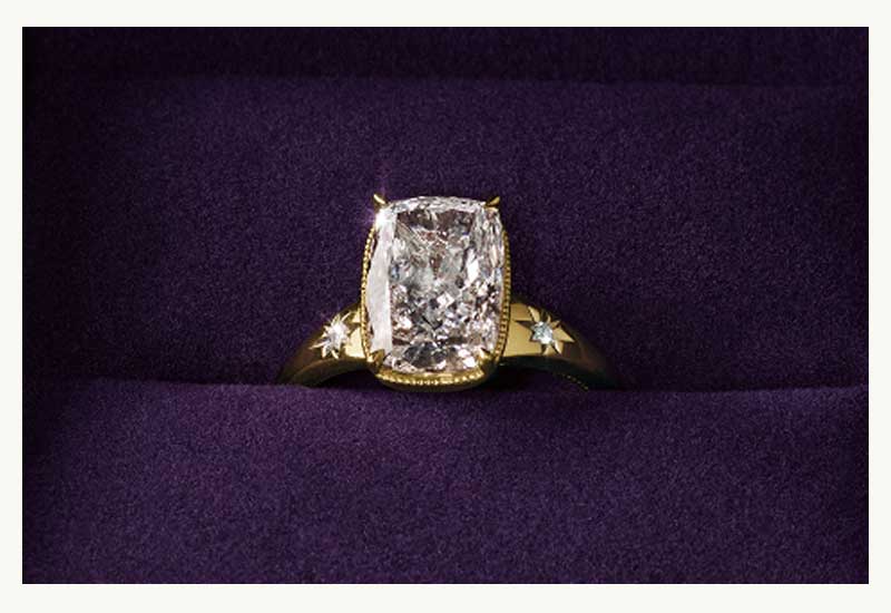 Gold cushion cut diamond ring with starburst engraved side stones, inside purple velvet ring box.