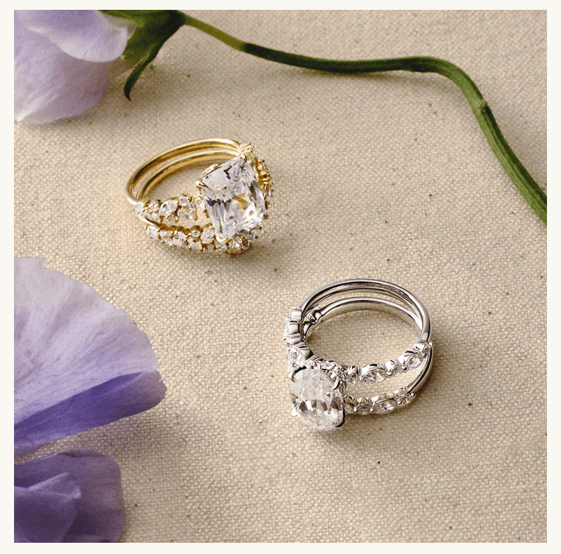 Diamond ring stacks, in yellow gold with radiant stone & white gold with oval stone, arranged next to flowers.