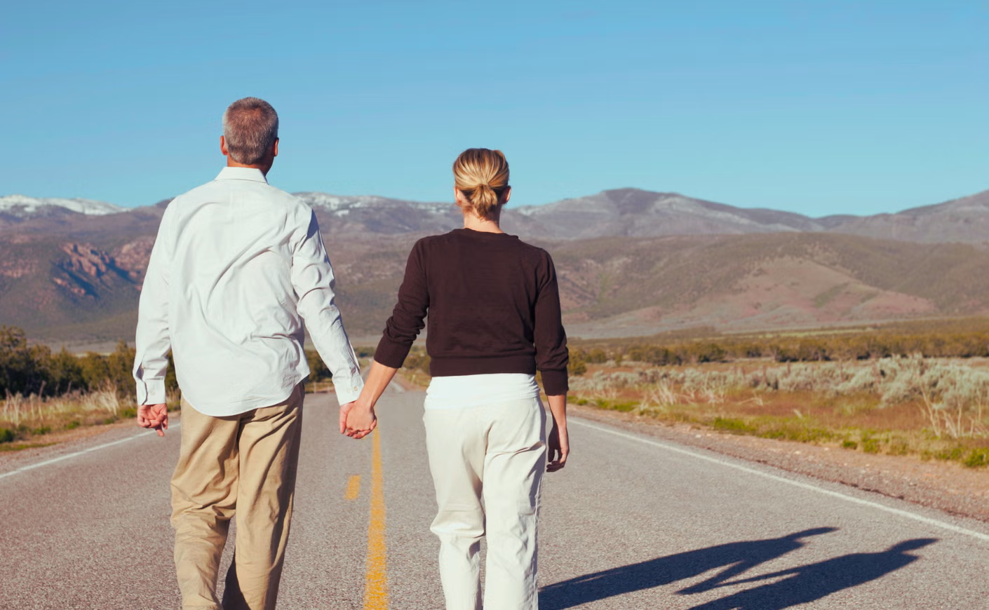A woman and a man holding hands while walking down a road leading to mountains