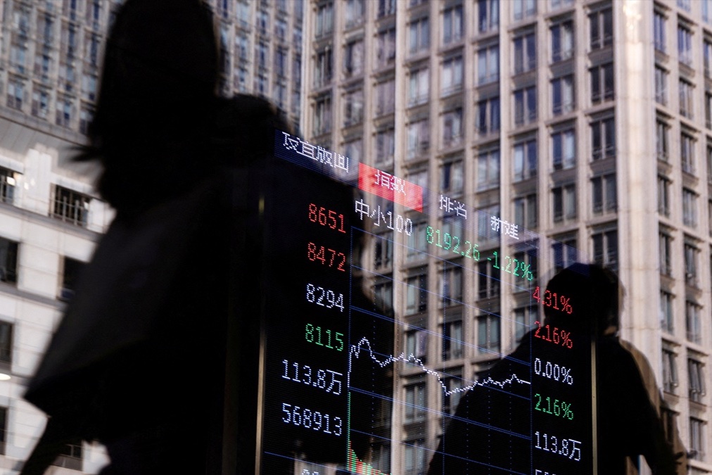 People walk past a brokerage house as an electronic board displays stock index information graph, in the Central Business District in Beijing, China, October 13, 2025.
