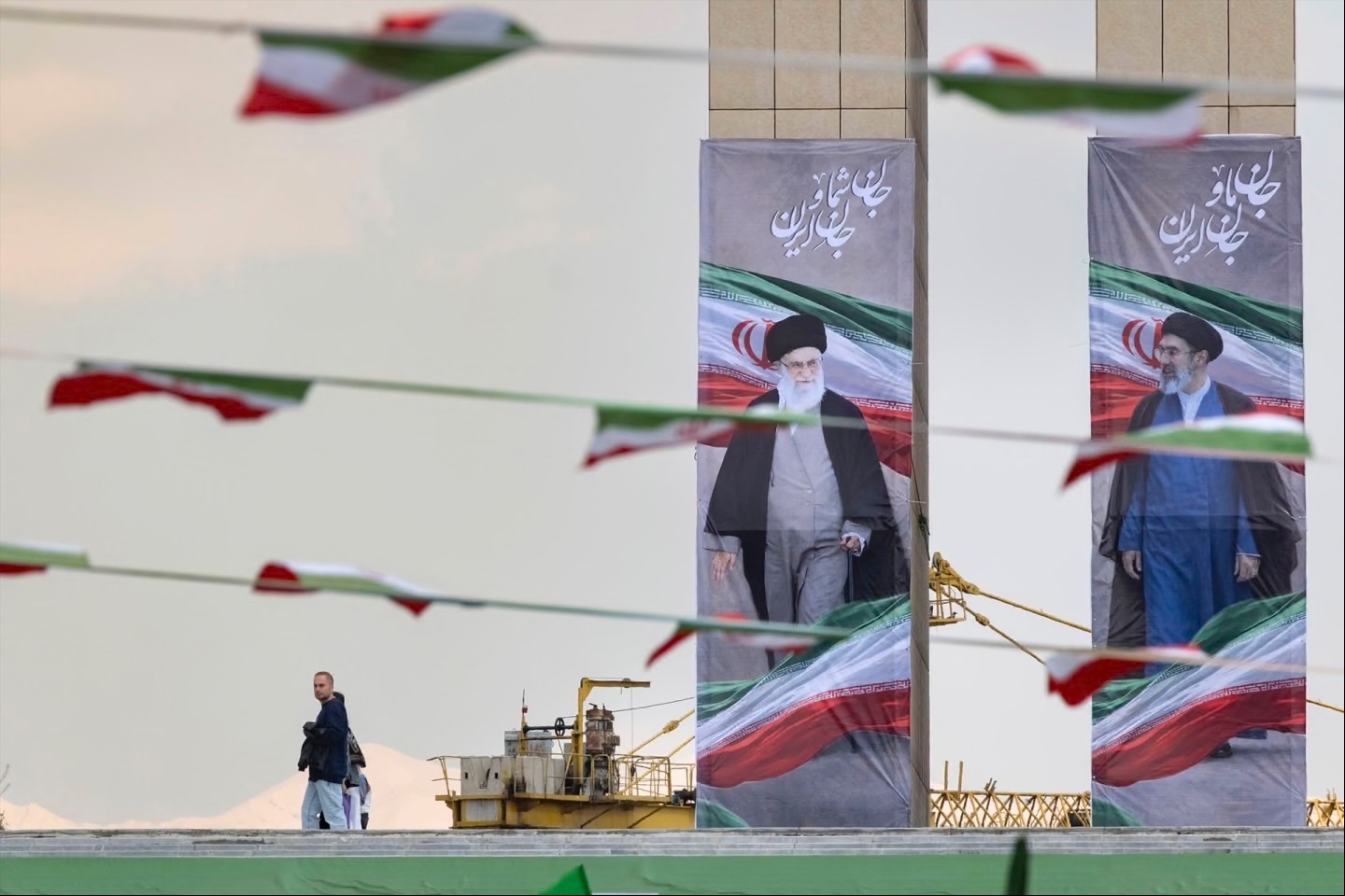 An Iranian man walks past the portraits of late Supreme Leader Ayatollah Ali Khamenei and the new Supreme Leader Ayatollah Mojtaba Khamenei during a flag ceremony marking Iran’s Islamic Republic National Day in the Abbasabad Cultural and Tourist Area in central Tehran, on April 1, 2026.