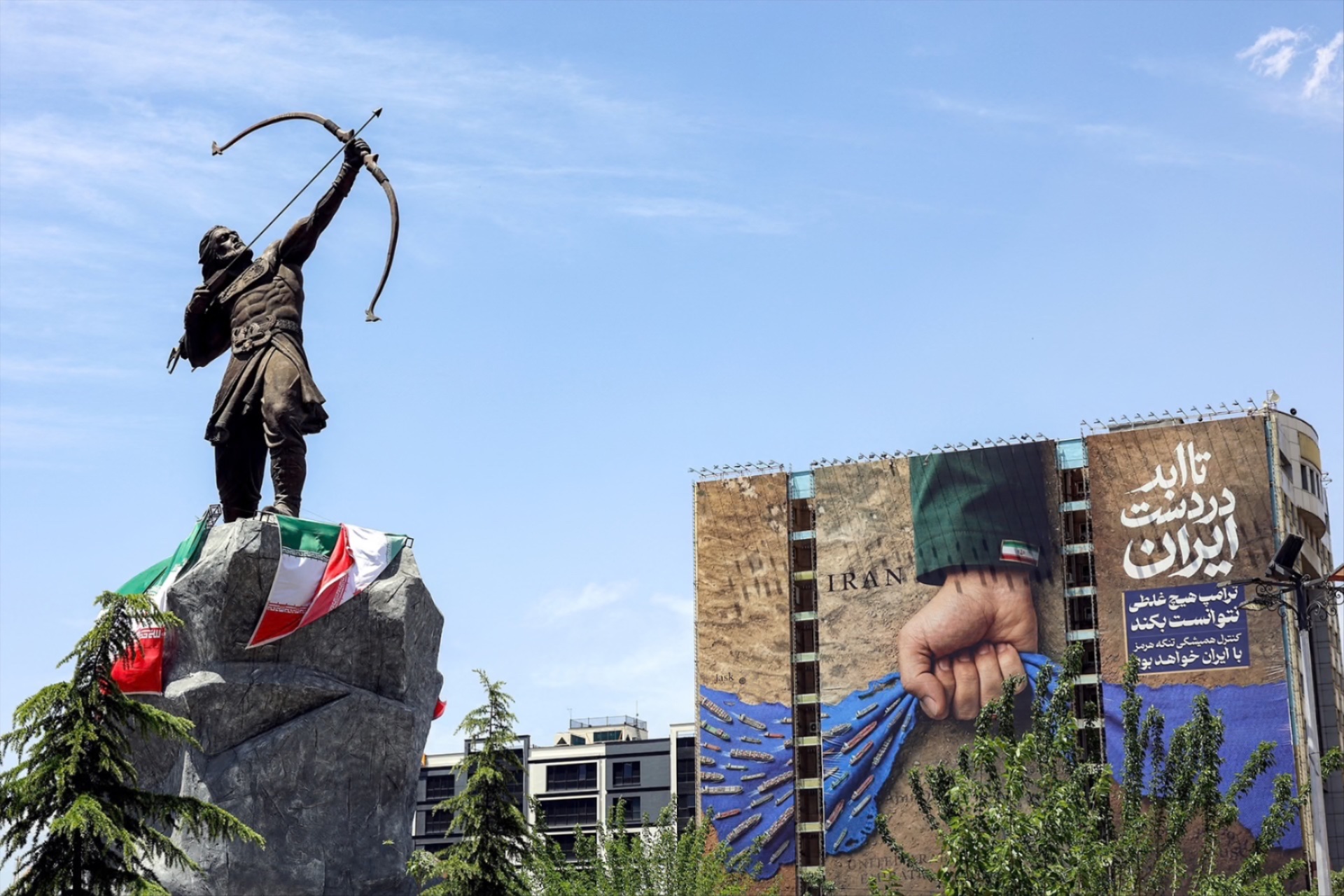 A statue depicting Persian mythical figure Arash the Archer is displayed near a billboard on the facade of a building depicting the Strait of Hormuz with a caption in Persian reading “Forever in Iran’s Hand”, at Vanak Square in Tehran on May 6, 2026.