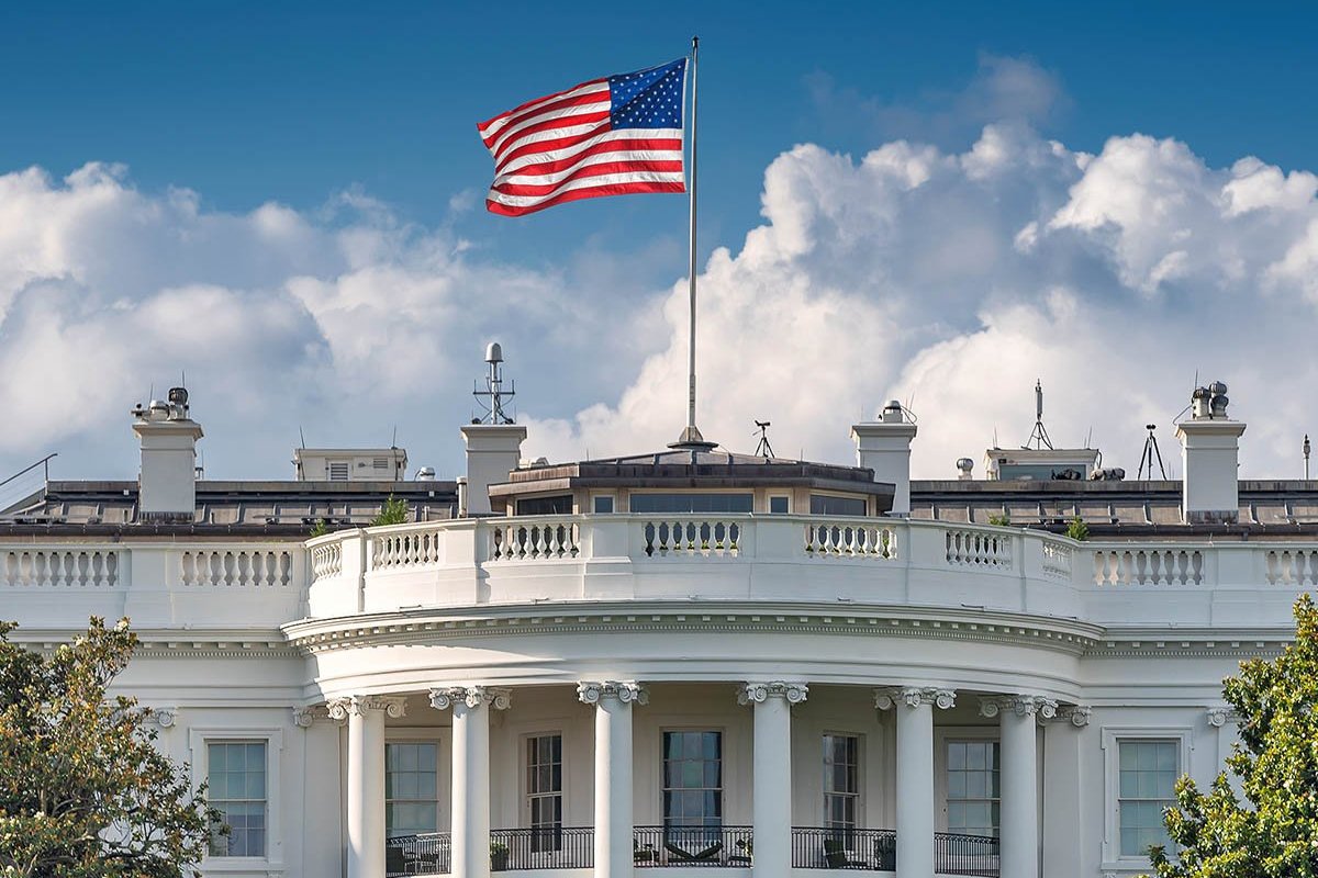 The White House with an American flag flying over it, in front of a background of blue sky and clouds. 