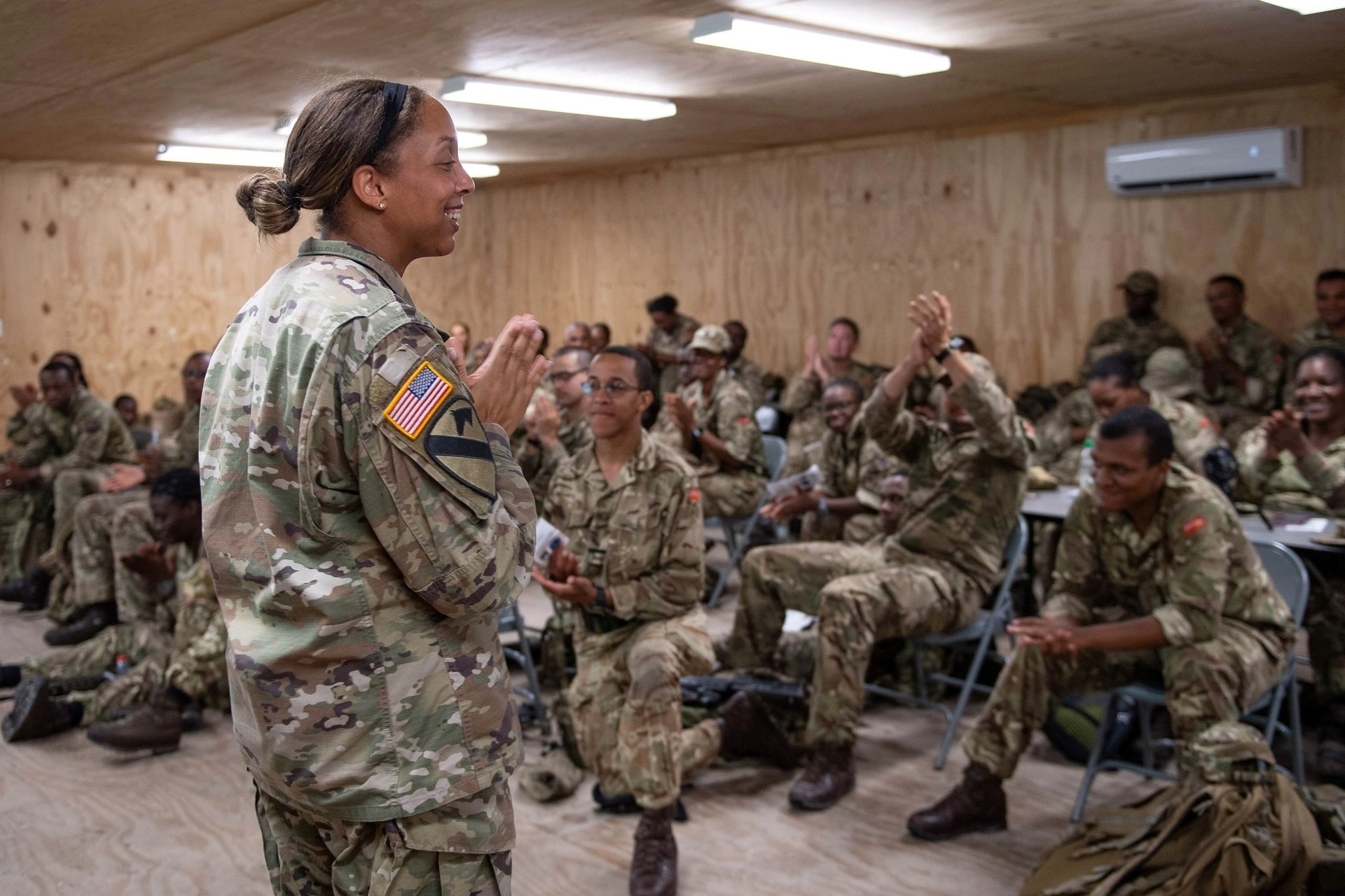 A Black female U.S. soldier stands before a group of soldiers in a wood-paneled room.
