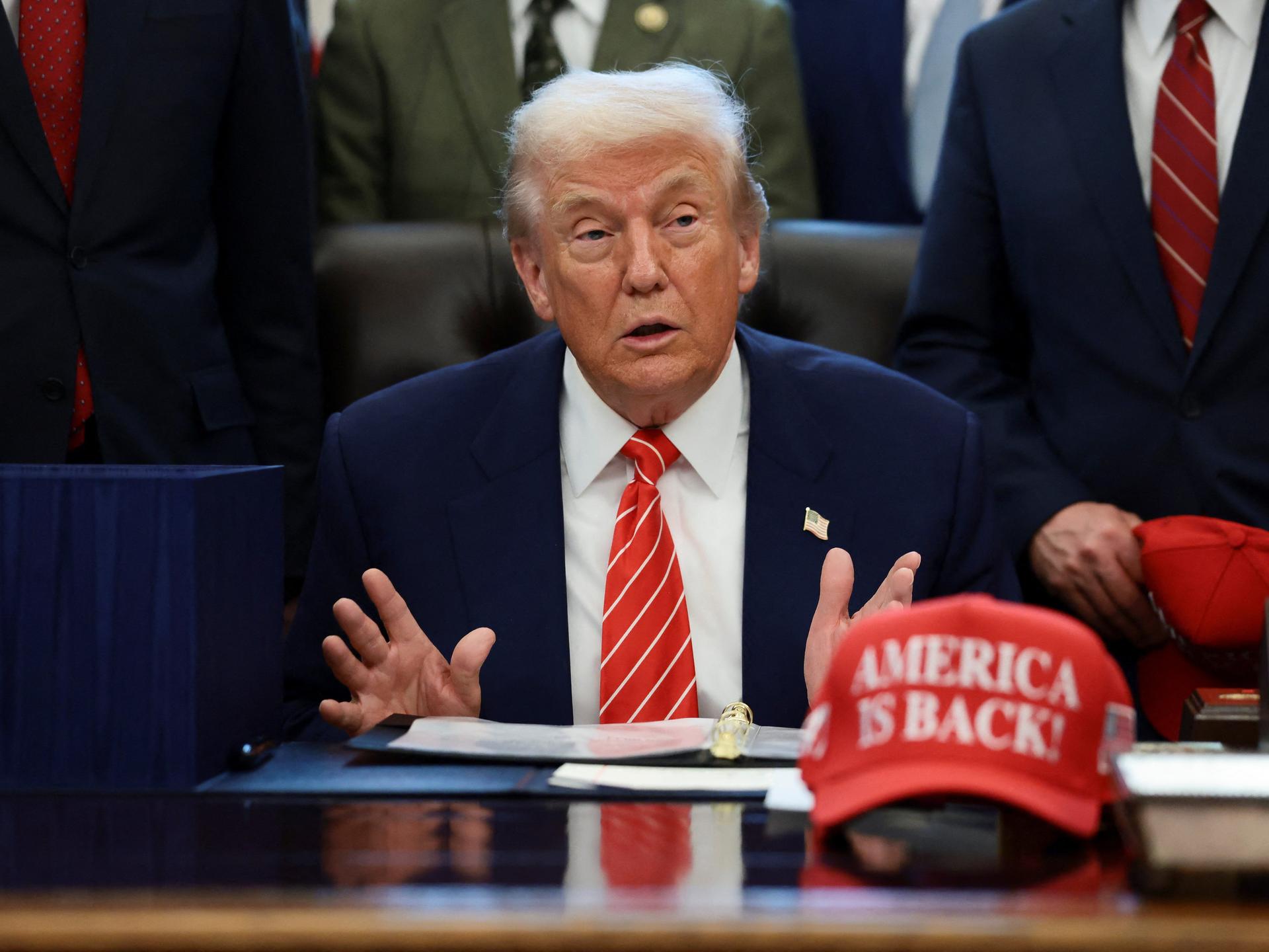 U.S. President Donald Trump sits at his desk, behind a hat that reads 