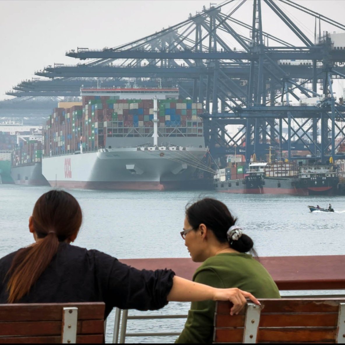 Women sit and talk on a waterfront bench facing large container ships 
