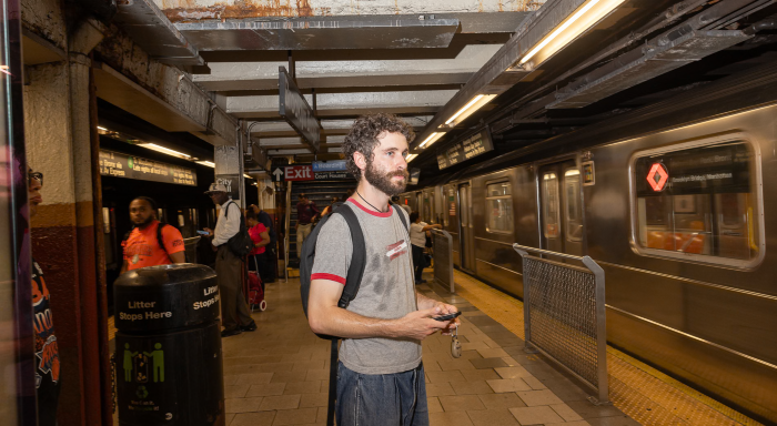 A man with curly brown hair, a mustache, and a beard wears a sweat-stained gray T-shirt. He stands on a subway platform with other people in the background.