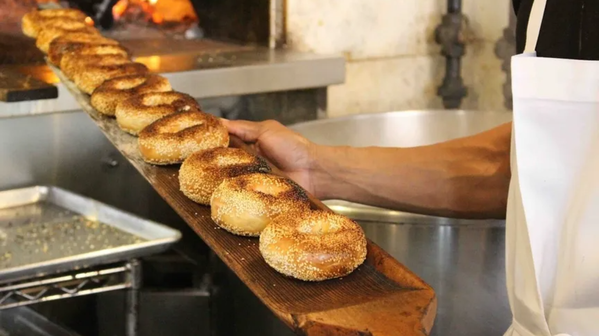 A muscled arm holds a long wooden paddle holding a row of sesame bagels.