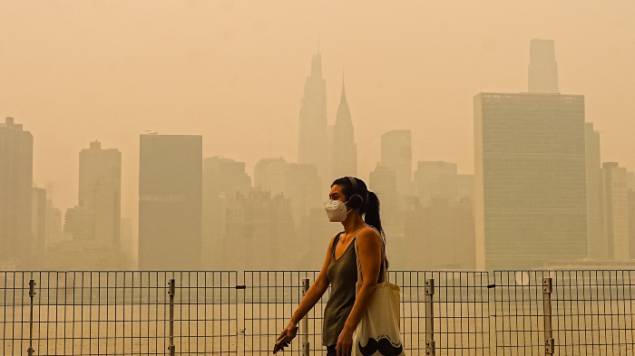A woman walks through a smoke-filled park wearing a mask, with the skyline of New York City in the background through the orange-tinged haze.