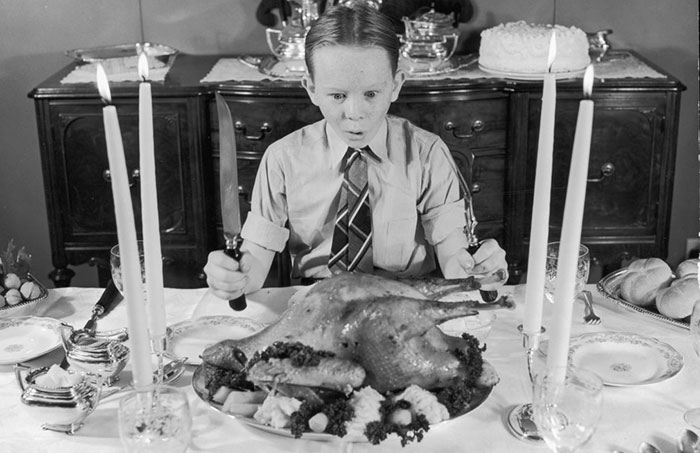 circa 1955, a black and white image of a redheaded boy, maybe 11 or so, sitting in shirt and tie at a table with a roast turkey in front of him, holding carving implements and looking very excited