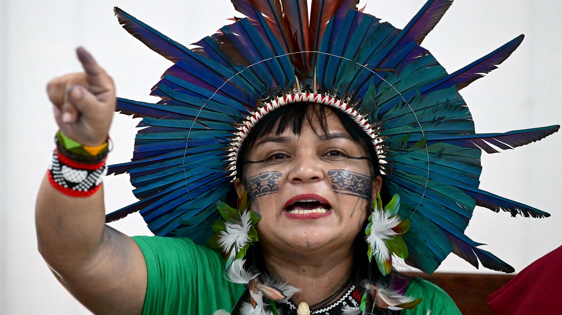 An indigenous leader wearing a blue feather headdress points her finger outward while speaking.  