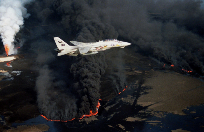 A pale gray military jet flies past a large black cloud of smoke from an oil well that has been set ablaze.