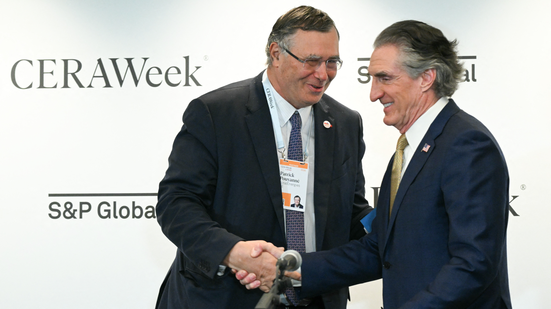 Two men wearing suits and ties handshake while smiling.