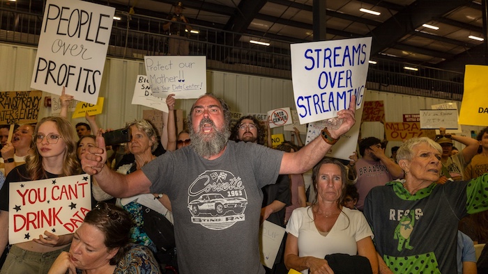 A group of angry people in a meeting room hold signs—people over profits, streams over streaming, you can't drink data—and yell at their local county leaders.