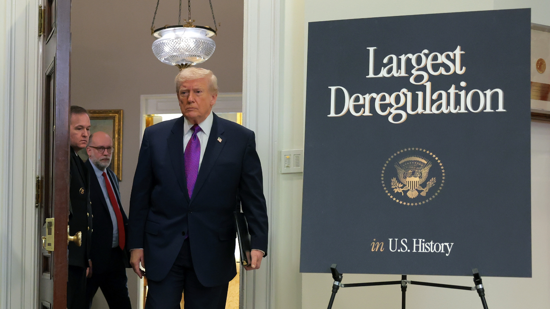 Donald Trump, wearing an oversized suit and purple tie, walks into a room next to a sign that says largest deregulation in U.S. History