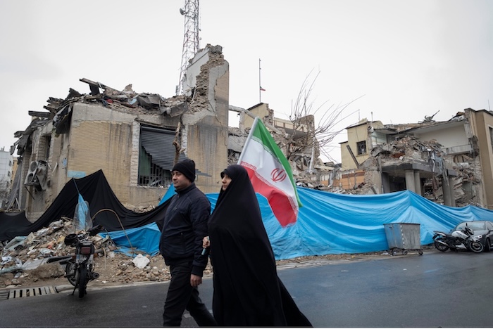 An Iranian couple carrying the national flag on March 13 walk past a police facility destroyed in an attack on Tehran. Morteza Nikoubazl/NurPhoto via AP