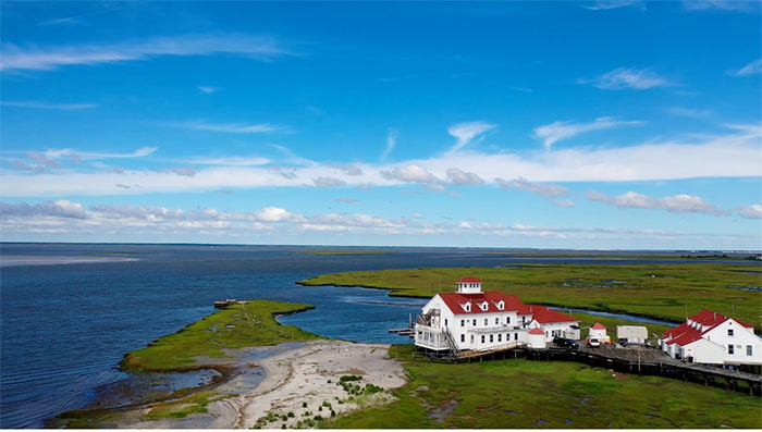 A wide shot of a gorgeous blue sky over the water, with some clouds, and a lush green wetland in the foreground with white house-like buildings with red roofs, very quaint.
