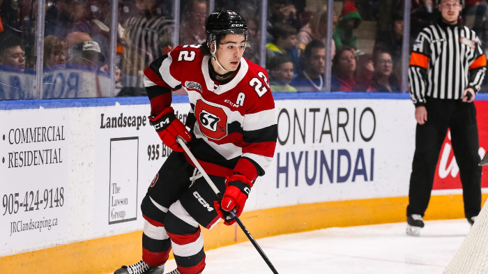 Matthew Mayich, a young Caucasian man in a hockey uniform, skates on the ice. He wears a red, black, and white jersey as #22 of the Ottawa 67's and holds a hockey stick. Dozens of spectators watch from behind glass.