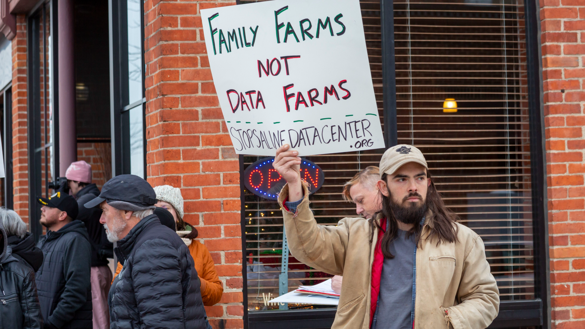 A person with a long brown beard and long brown hair holds a sign that says family farms not data farms