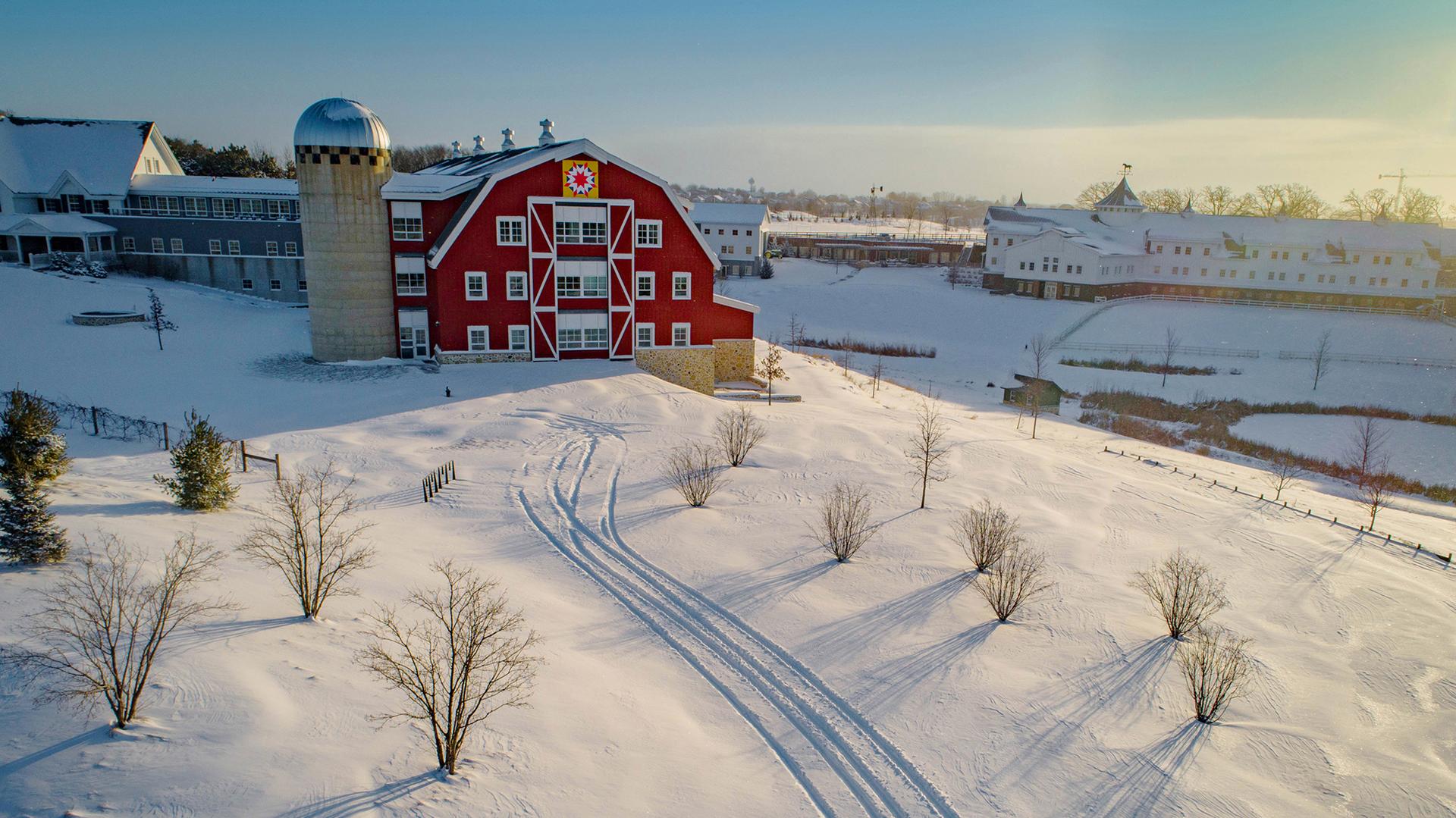 A snowy field in front of a red barn, illuminated by sun.